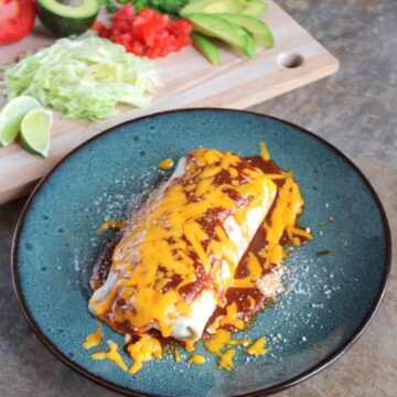 Lightened-Up Smothered Burritos: a burrito on a blue, ceramic plate. In the background sits a cutting board with various garnishes, including sliced avocados, sliced scallions, lime wedges, and chopped cilantro.