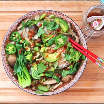 Overhead view of a bowl of Instant Pot Vegetable Pho Soup, loaded up with herbs and garnishes, with a red pair of chopsticks. The bowl sits next to a jar of vegan nuoc cham.