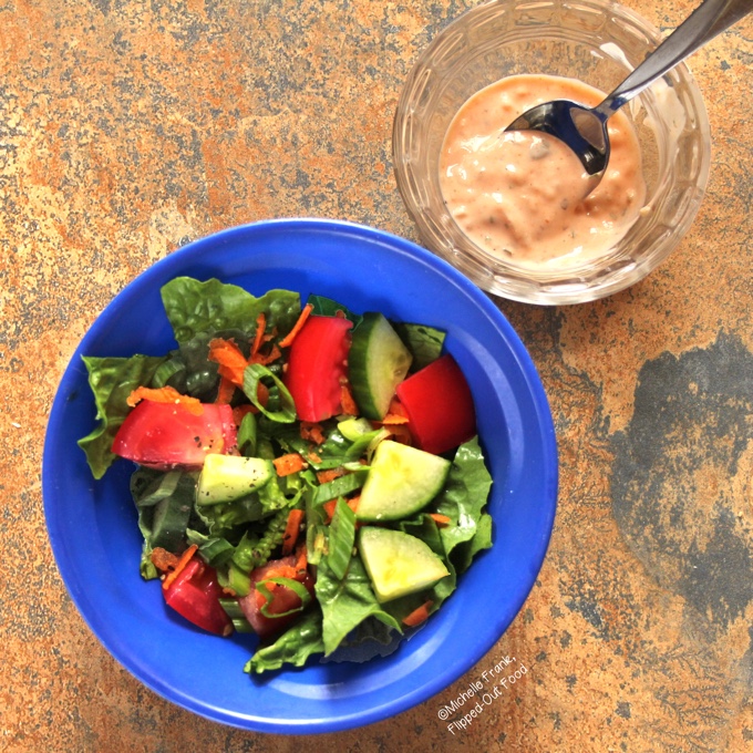 An individual serving of Meal-Prep House Salad & Homemade Thousand Island Dressing in a blue bowl. The salad is piled with tomatoes, sliced cucumbers, scallions, and grated carrots. The dressing is on the side in a clear ramekin.