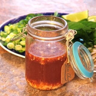 Quick Nuoc Cham Sauce: a side view of a mason jar full of sauce sitting in front of a platter of herbs, lime wedges, and sliced scallions and jalapenos.