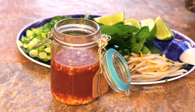 Quick Nuoc Cham Sauce: a side view of a mason jar full of sauce sitting in front of a platter of herbs, lime wedges, and sliced scallions and jalapenos.