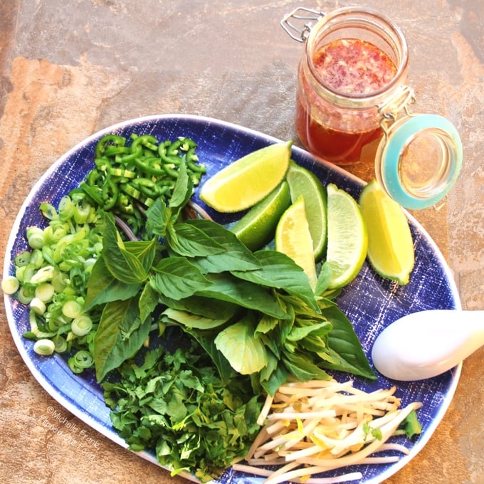 Top view of a jar of Quick Nuoc Cham Sauce sitting next to a blue platter loaded with condiments for pho, including sliced scallions and jalapenos, lime wedges, mung bean sprouts, Thai basil, and chopped cilantro.