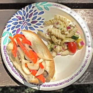 Grilled Italian Sausage Sandwiches with Pepper-Onion Foil Packs on a decorative paper plate with Greek Pasta Salad. The plate is sitting on a rustic picnic table located at Devil's Lake State Park in Wisconsin.