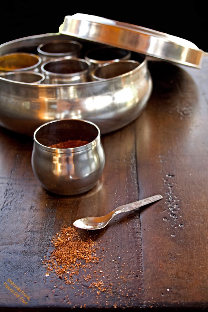 5-Ingredient Homemade Taco Seasoning scattered on a tabletop in front of a tiny spoon. Behind sits a metal cup and an Indian spice box.