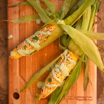 Elote: Mexican Street Corn with Jalapeno-Cilantro Crema. 2 ears sit on a wooden cutting board, with husks flying, drizzled with crema and sprinkled with cilantro.