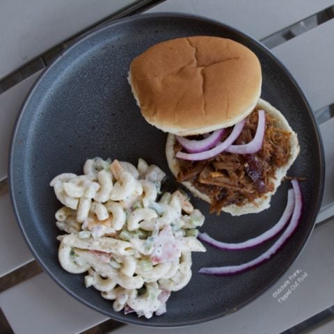 Creamy Pasta Salad: a serving on a grey plate next to a pulled pork sandwich and sliced red onion.