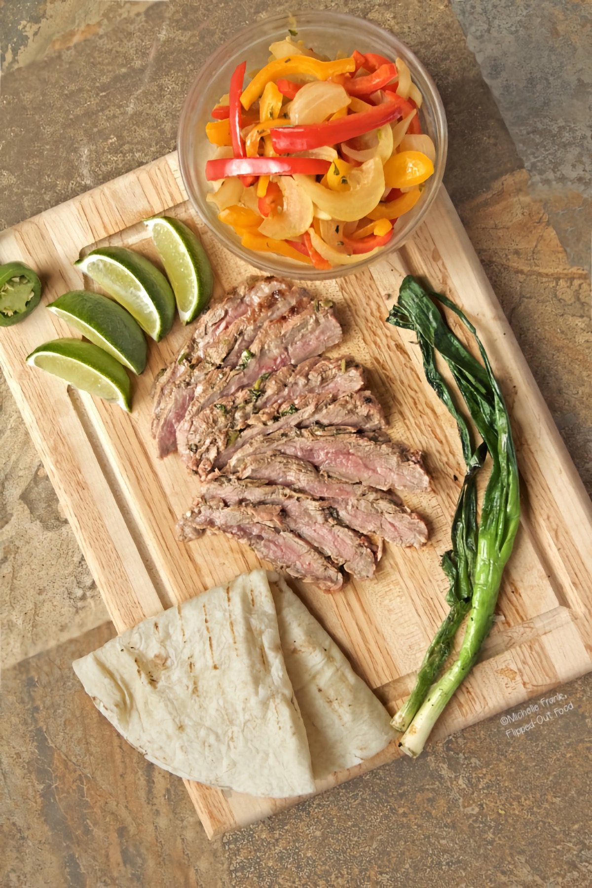 Top view of carne asada with fajita vegetables arranged on a cutting board with a folded flour tortilla, grilled scallions, lime wedges, and the cut end of a jalapeno pepper.