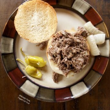 Instant Pot Mississippi Pot Roast Sandwiches: a single sandwich on a plate with onions and pepperoncini peppers. The top bun is set on the side.