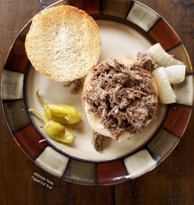 Instant Pot Mississippi Pot Roast Sandwiches: a single sandwich on a plate with onions and pepperoncini peppers. The top bun is set on the side.