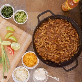One-Skillet Taco Pasta in its skillet, sitting beside a bottle of cholula. In the foreground are several garnishes, including choppoed tomatoes and scallions, shredded cheese, and lime wedges.