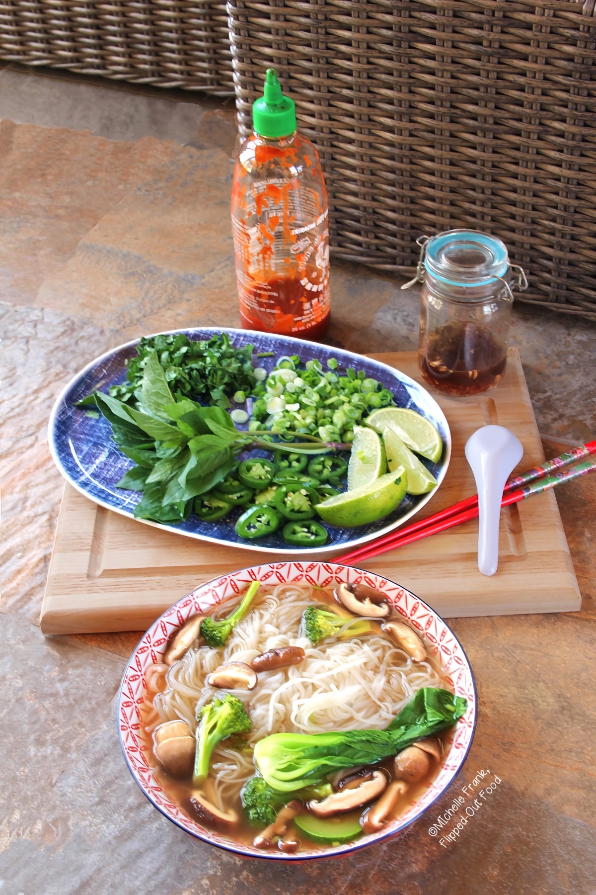 Easy Soups for Fall: Instant-Pot Vegetable Pho served in a red and white bowl. Behind the bowl are a platter with garnishes, a jar of vegan nuoc cham, a bottle of sriracha, chopsticks, and a spoon.