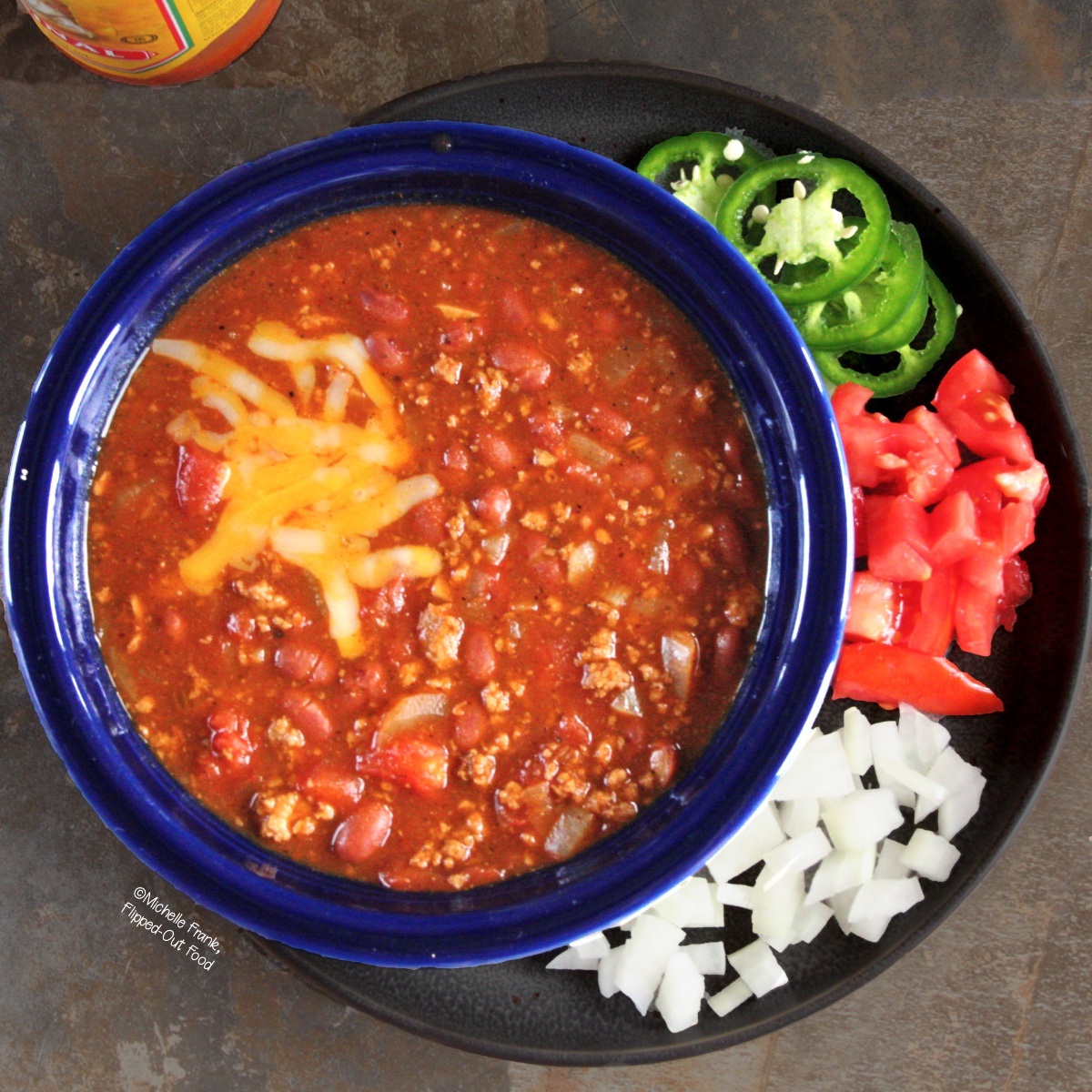 Easy Soups for Fall: Easy Turkey Chili, served in a blue bowl and topped with shredded cheese. The plate underneath the bowl holds garnishes including sliced jalapenos and chopped tomatoes and onions.