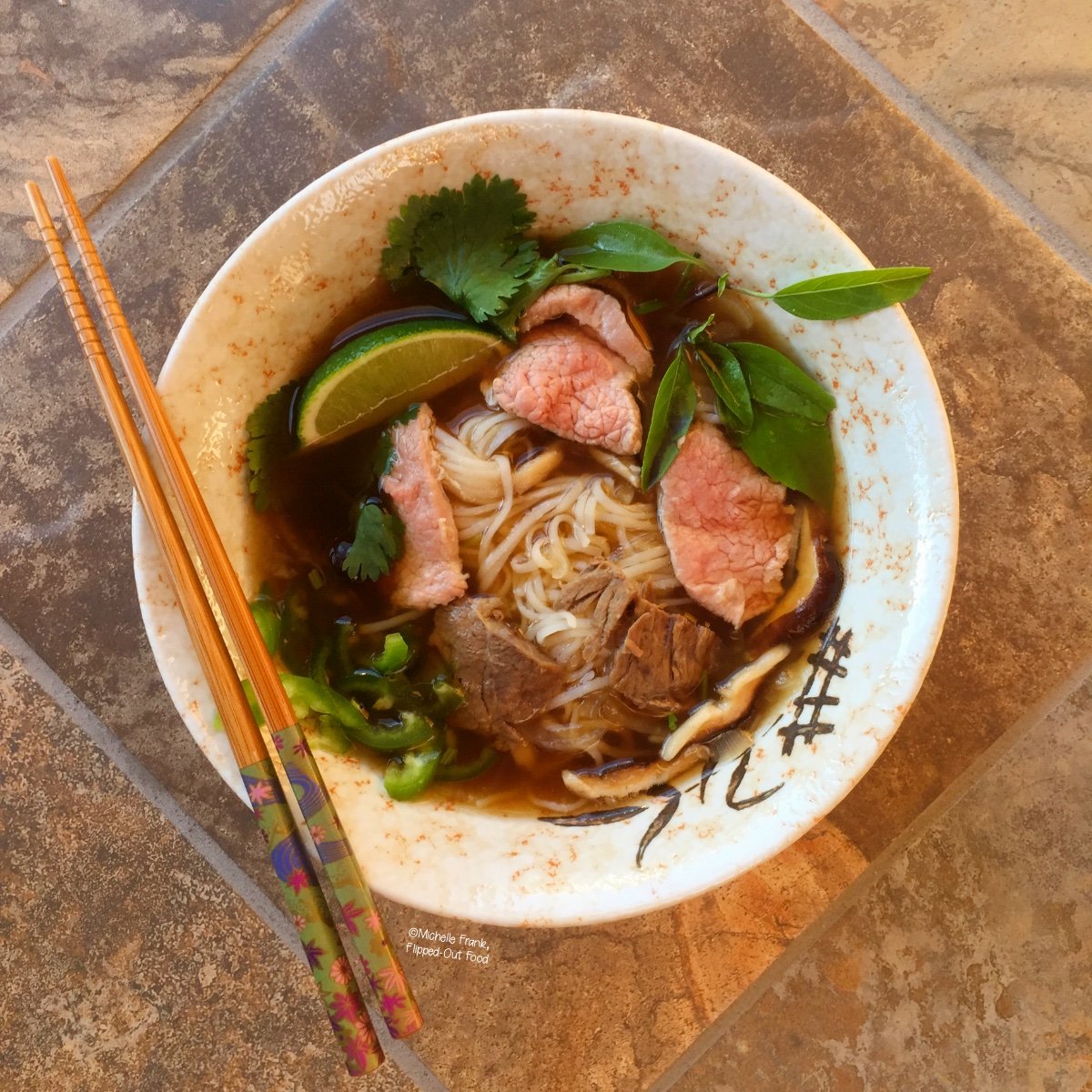 A bowl of Beef Pho Noodle Soup, garnished with herbs and a lime wedge. A pair of chopsticks sits on top of the bowl.