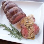 Roast Beef Tenderloin with Green Peppercorn Sauce, the roast on a white platter with two sliced medallions covered in sacue.