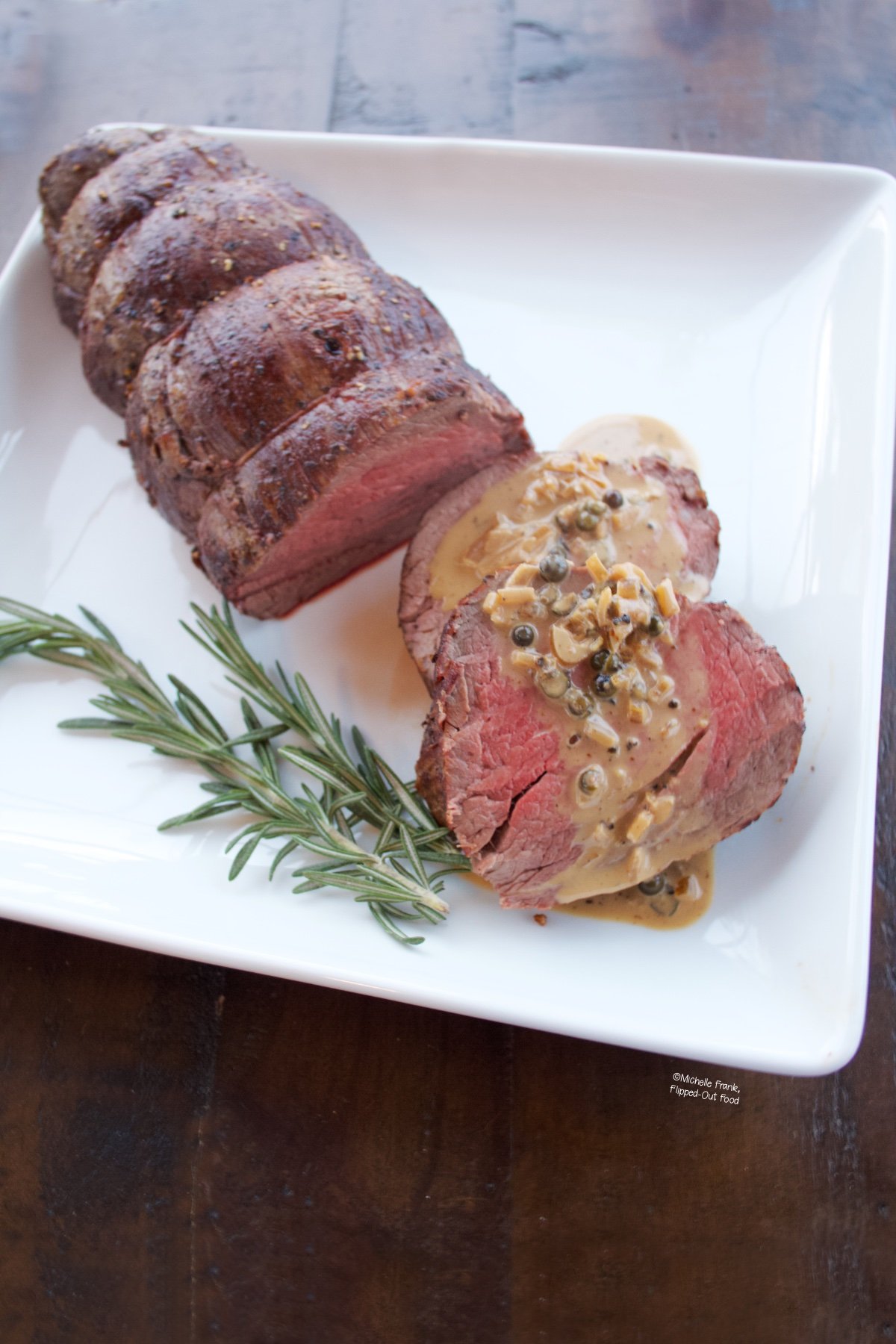 two slices of medium rare tenderloin form a whole tender on a white plate with peppercorn sauce on top of them.