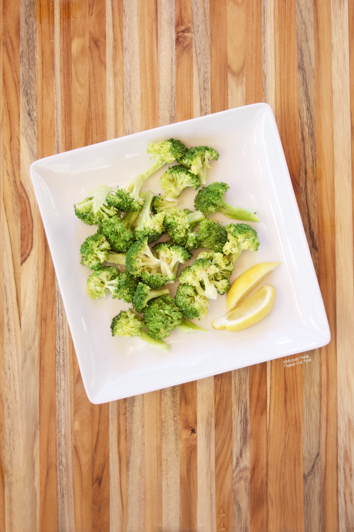 overhead image of cooked microwave broccoli on a white plate with lemon wedges, sitting atop a teak cutting board.