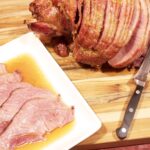 A platter of Orange-Bourbon Glazed Ham slices on a white platter with pan juices. Next to it sits the rest of the ham on a cutting board with a carving knife.