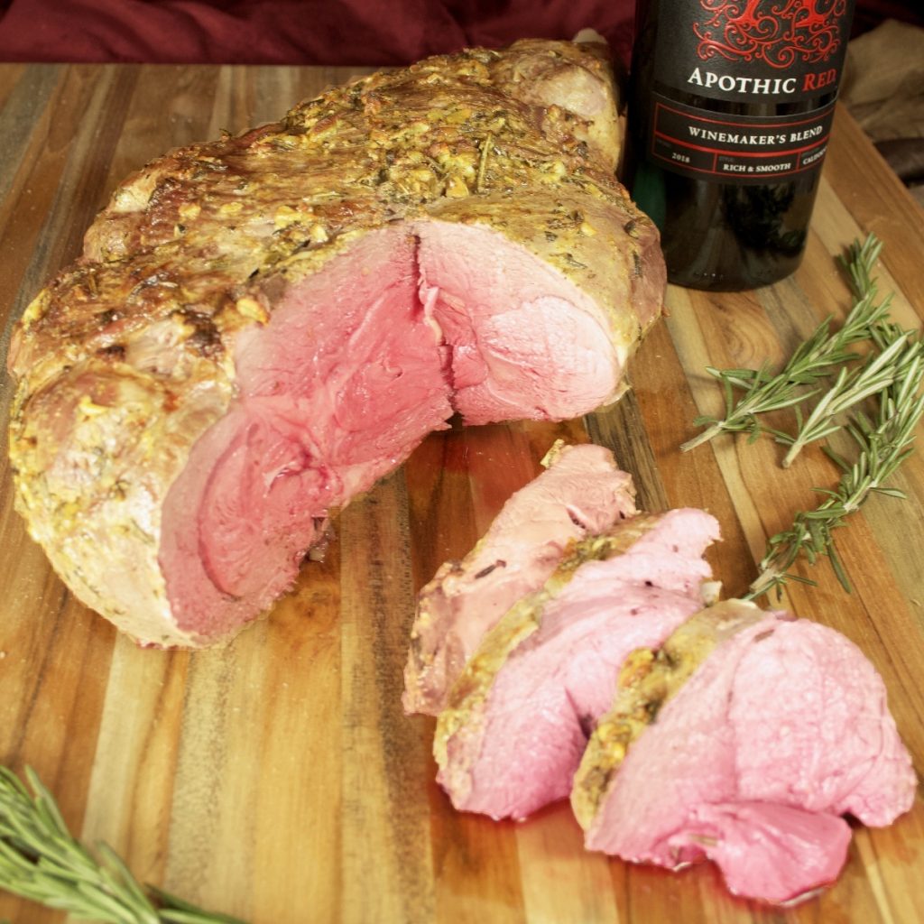 Side view of the partially sliced roast leg of lamb on a cutting board with rosemary sprigs and a bottle of wine in the background.