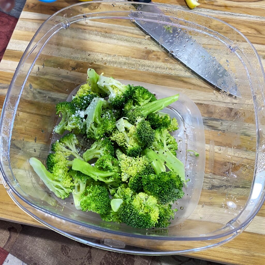The cooked microwave broccoli in a bowl with Parmesan cheese, salt, pepper, and lemon juice, ready to be tossed together.