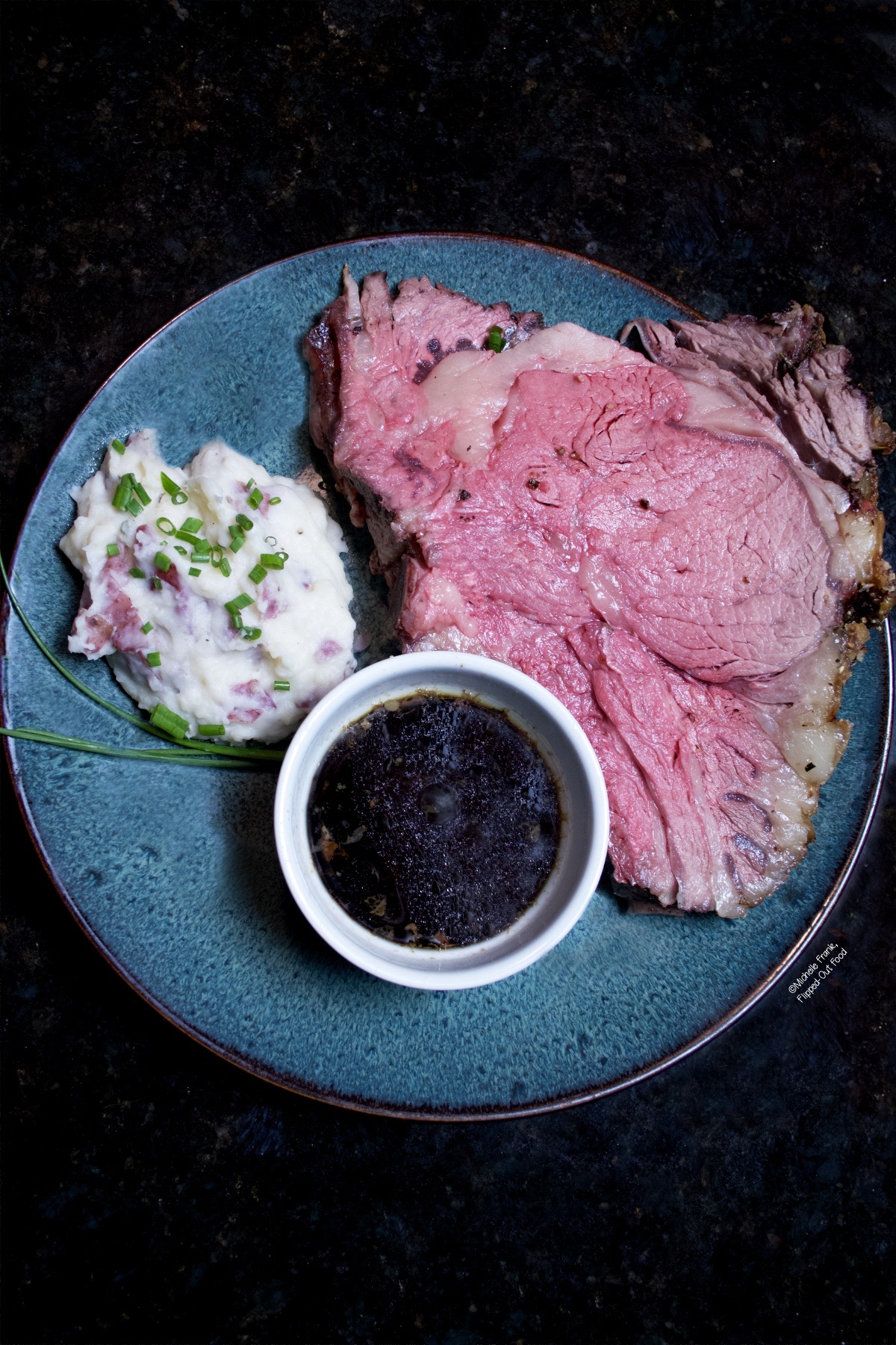 Overhead view of a beautiful slice of prime rib cooked to medium rare on a blue plate served with mashed potatoes and homemade au jus.