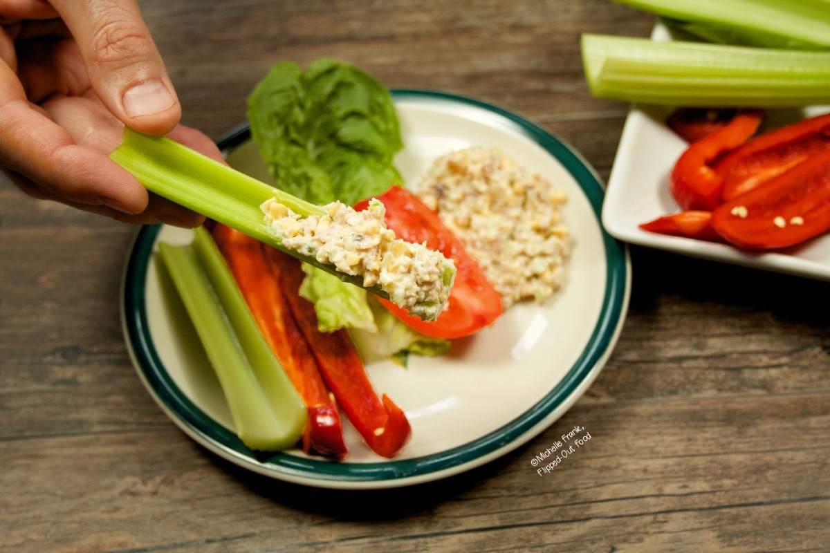 Close up view of a person holding a celery stick loaded with keto ham salad. In the background is a plate with sliced vegetables.