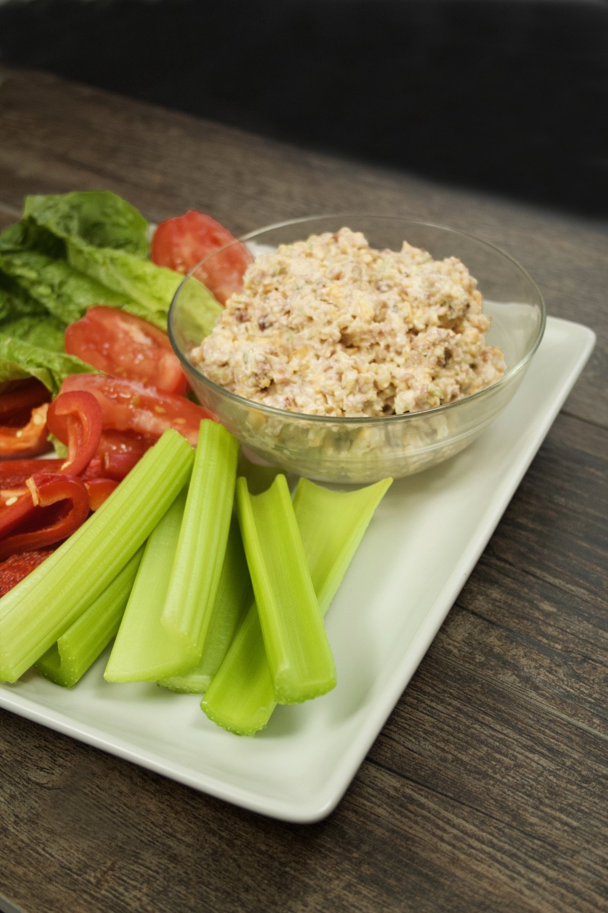 Side view of a serving bowl with Keto Ham Spread set on a plate with sliced vegetables and celery sticks.