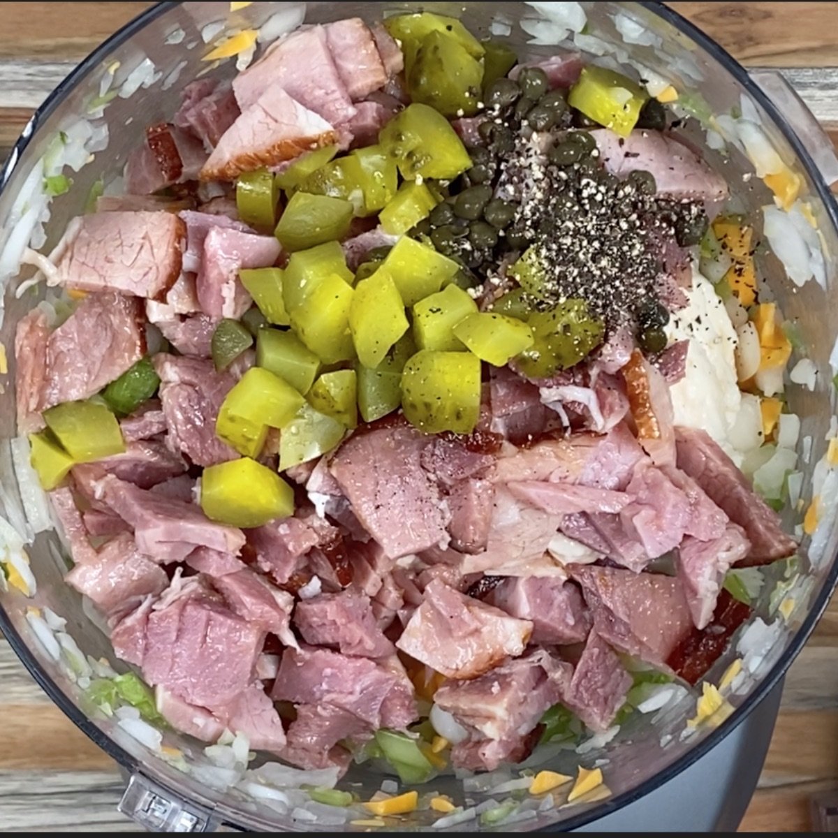 Overhead view of ham, pickles, capers, salt, pepper, mayonnaise, and mustard added to the food processor about to be blended.