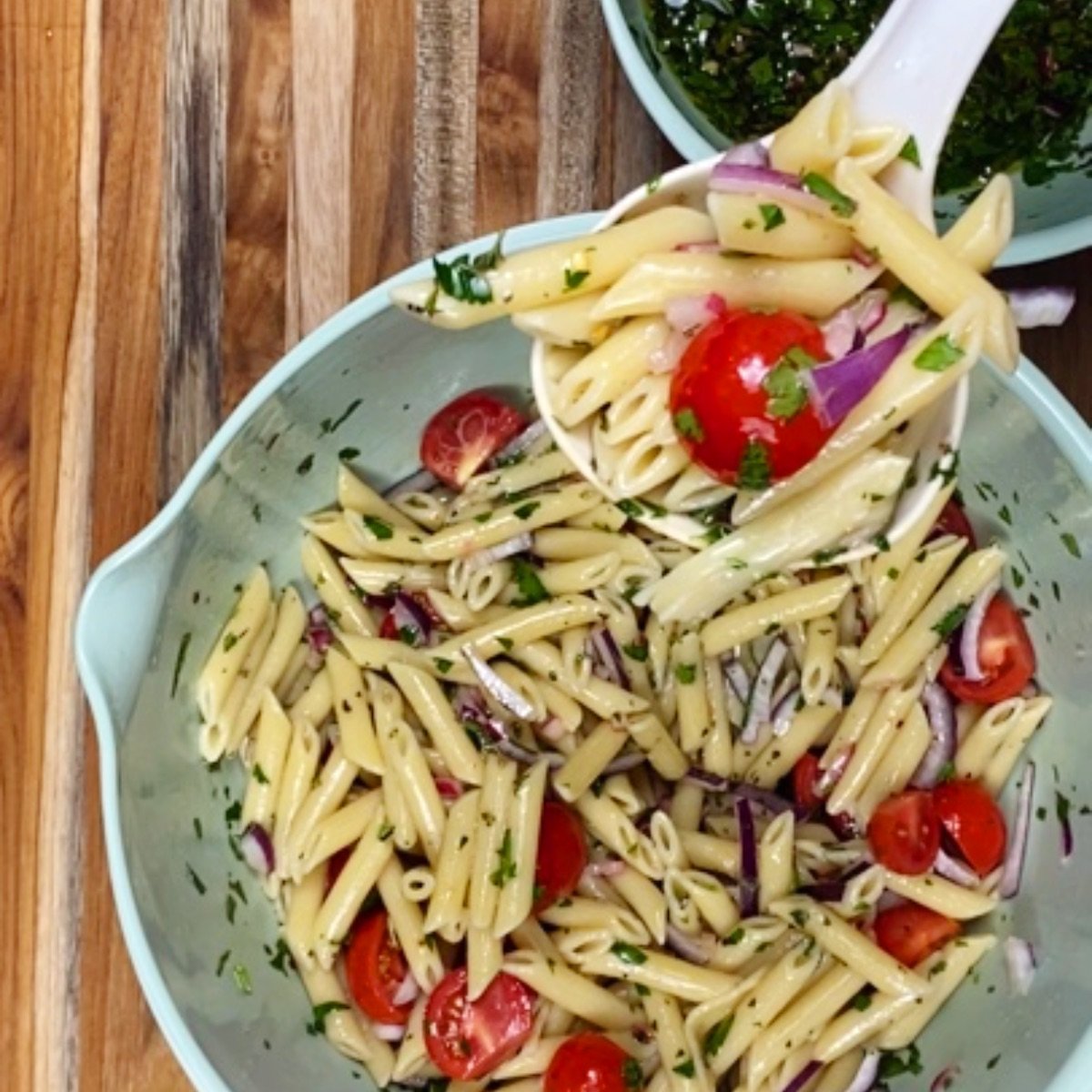 A serving spoon of chimichurri pasta salad being raised toward the camera, with the mixing bowl full of pasta salad underneath.