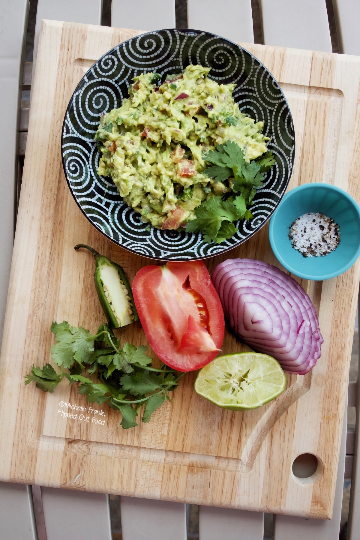 Easy Cutting-Board Guacamole in a black bowl set atop a wooden cutting board. Next to the bowl sits a ramekin with salt and pepper, a cut red onion, squeezed lime, cut tomato, cut jalapeno, and a sprig of cilantro.