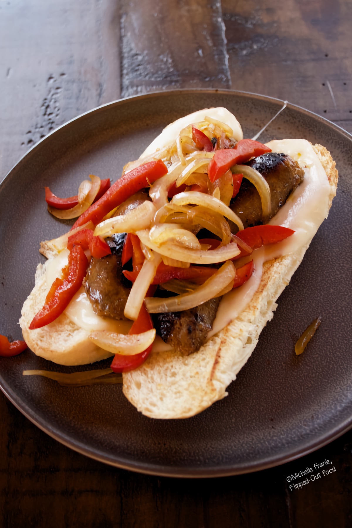 A Grilled Italian Sausage Sandwich with peppers and onions on a grey plate sitting atop a dark wood table.