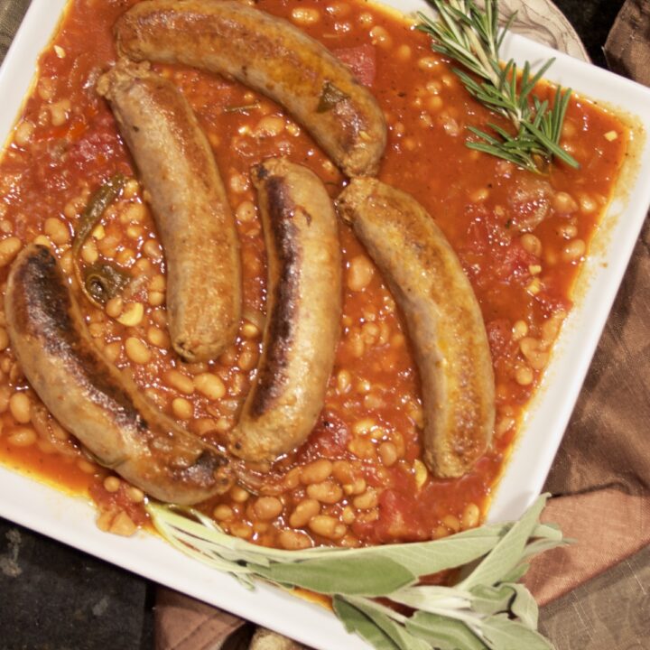 Italian Sausage and Beans (Salsiccie e Fagioli) on a white serving platter garnished with sprigs of sage and rosemary.