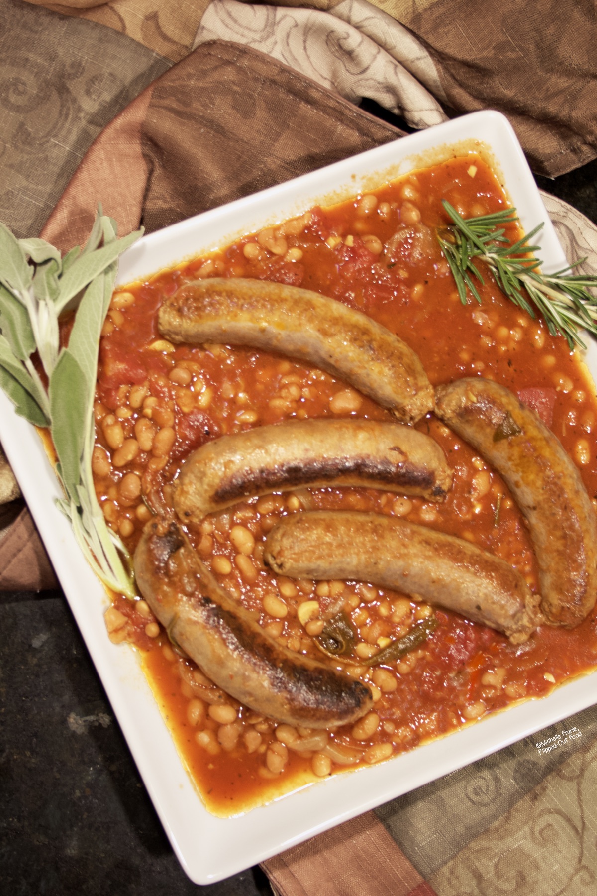 Italian Sausage and Beans (Salsiccie e Fagioli) on a white serving platter garnished with sprigs of sage and rosemary. 