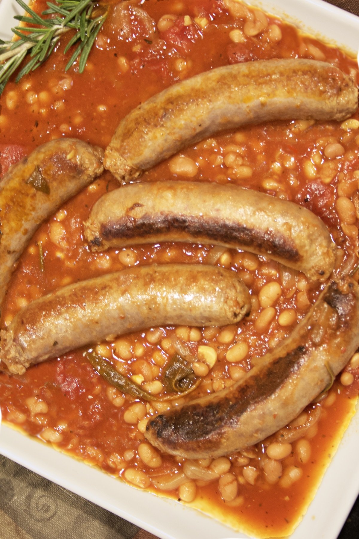 A close-up of a platter with Italian Sausage and Beans, garnished with a sprig of rosemary.