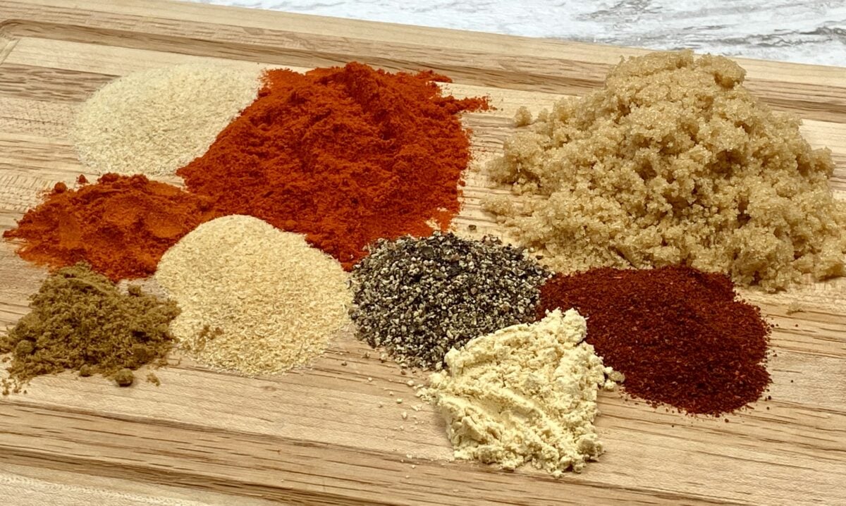 Side view of all the pork butt rub ingredients in piles on a small cutting board. Going roughly clockwise from lower left, the ingredients are Ingredients for my pork butt rub, including cumin, garlic powder, cayenne, onion powder, sweet paprika, ground black pepper, brown sugar, chili powder, and ground mustard. 