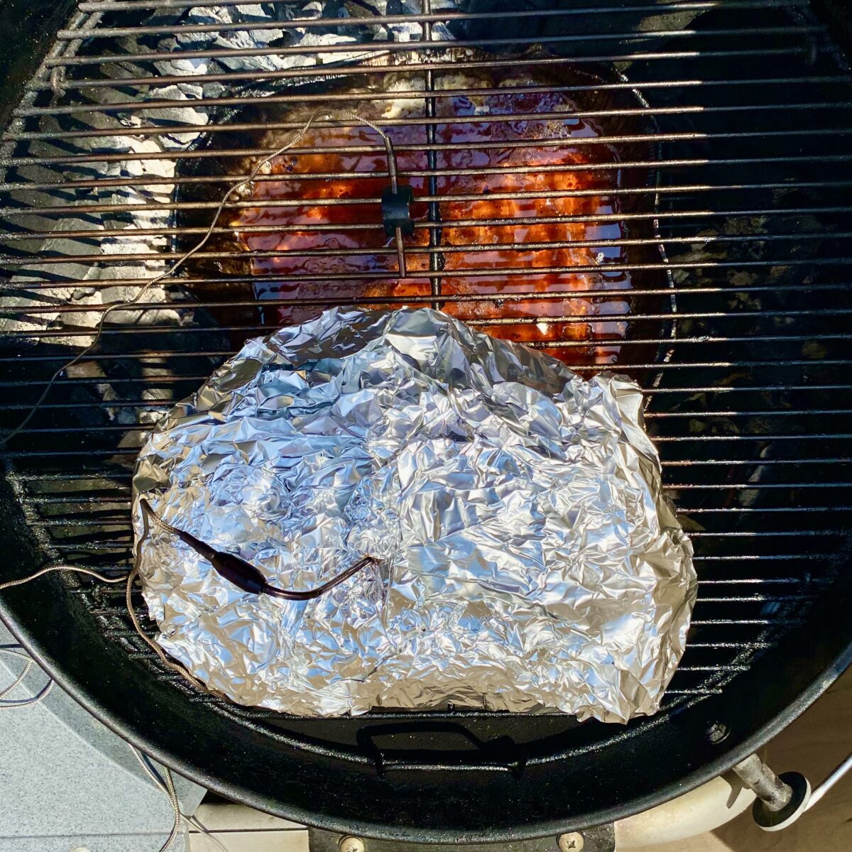 Top view of double wrapped pork butt on charcoal grill with meat thermometer inserted and properly positioned on the charcoal grill.