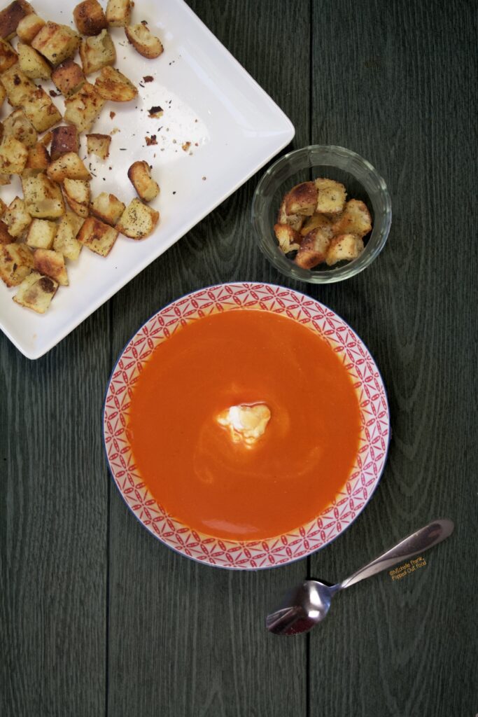 A bowl of Fresh tomato soup with a dollop of ricotta, sitting next to a platter and a ramekin with freshly made stovetop croutons.