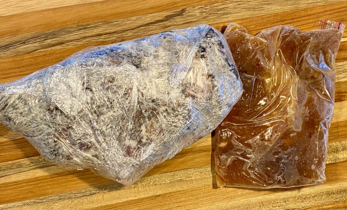 Frozen brisket and broth wrapped in plastic positioned on a cutting board.