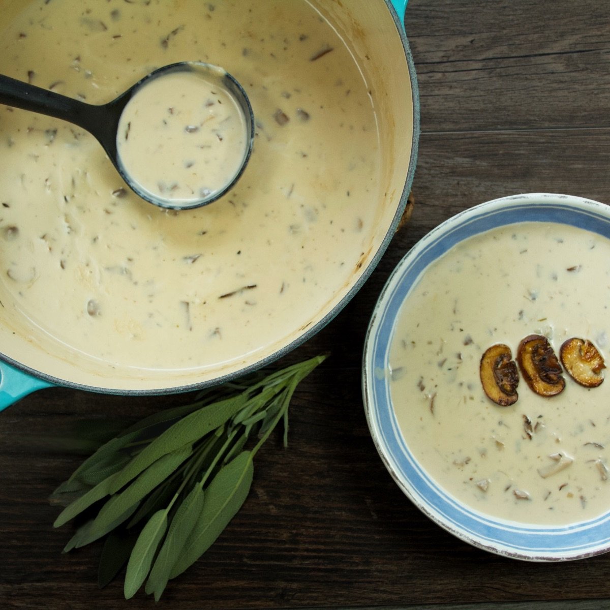 Top view of a dutch oven full of homemade cream of mushroom soup with a full ladle inside it. This sits next to a blue and white bowl with a serving of the soup. The soup has been topped with three golden-brown mushroom slices.