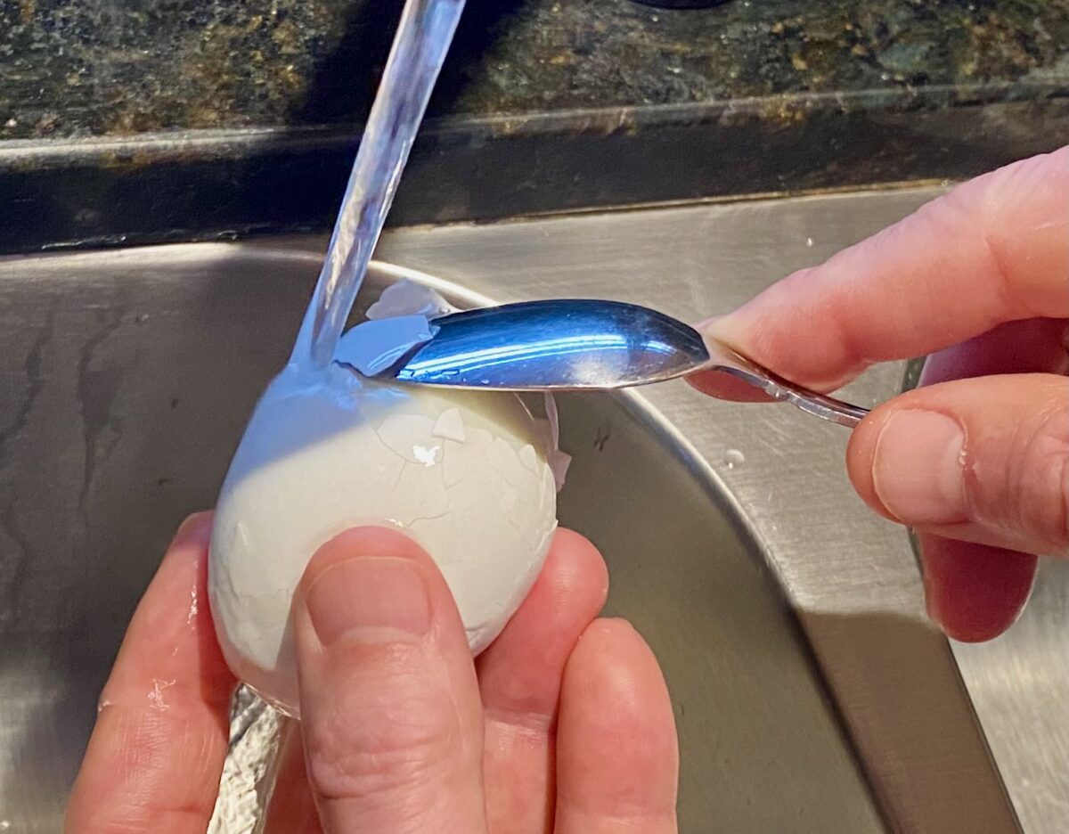 Side view of a partially peeled hard boiled egg with a small spoon pried between the shell and the egg white while under running water in a sink.