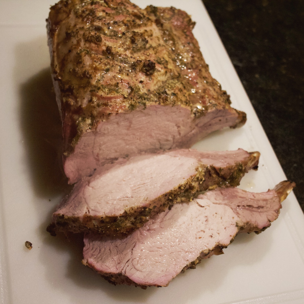 Side view of fully cooked rack of pork with two chops sliced off on white cutting board.
