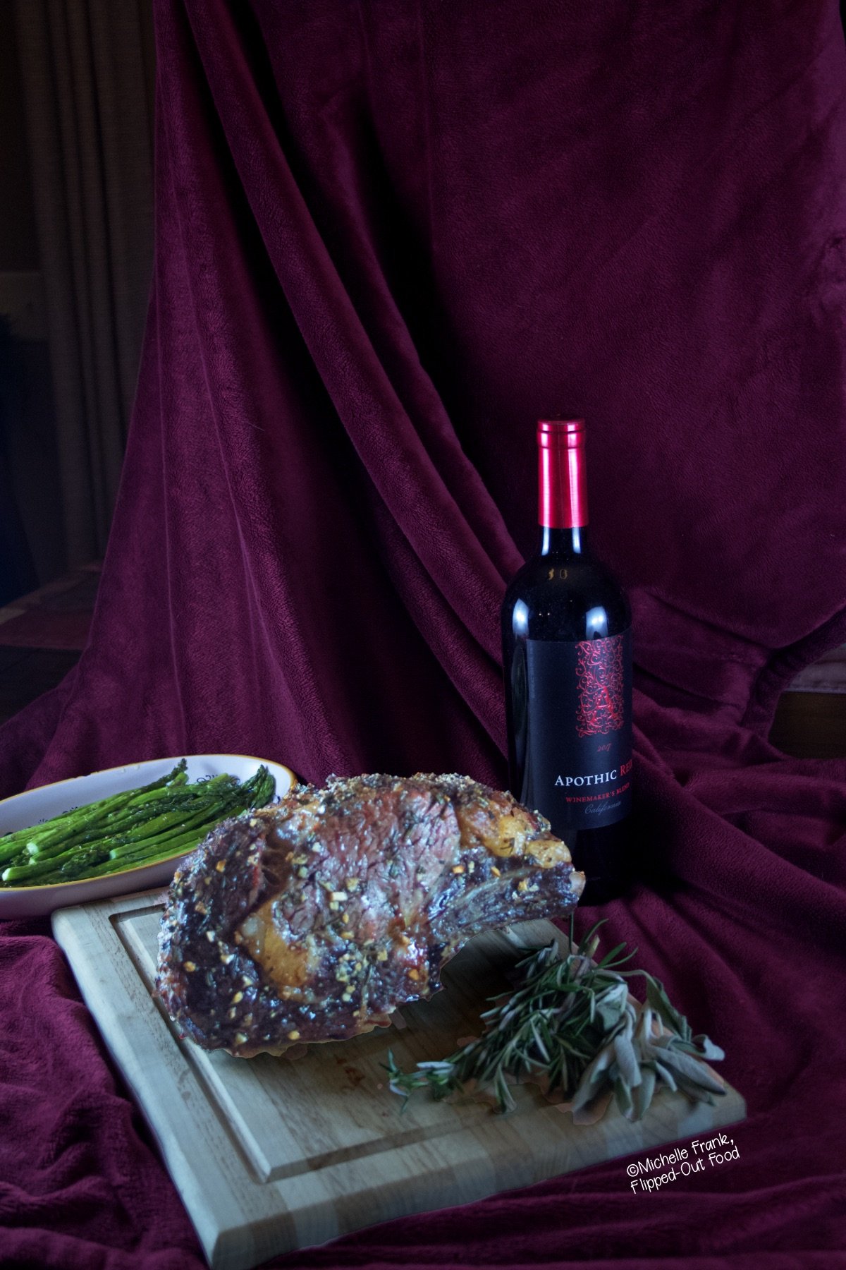 Date Night Prime Rib roast resting on a cutting board surrounded by herbs (rosemary and sage). In the background is a dish of roasted asparagus and a bottle of red wine.