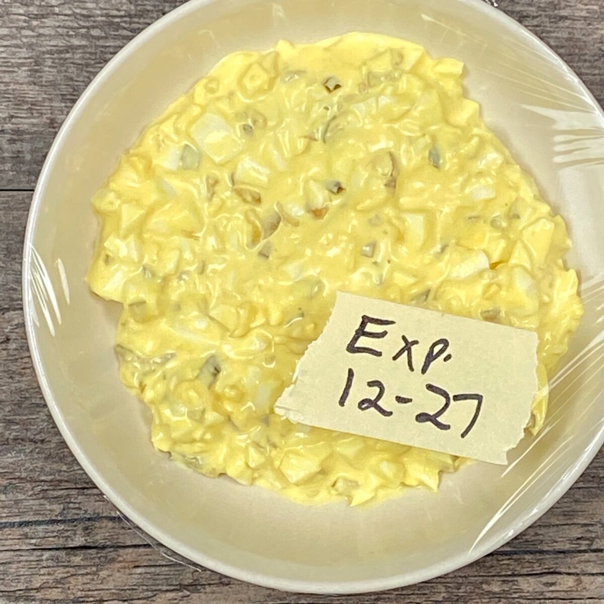 A bowl of egg salad, covered with plastic wrap, and labeled with the expiration date.