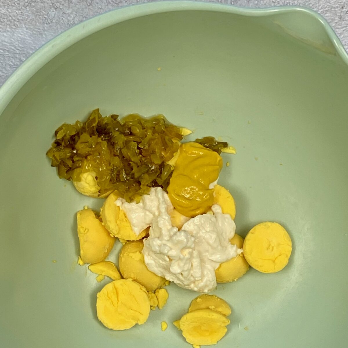 Overhead view of hard boiled egg yolks, mayonnaise, mustard and relish in a green bowl about to be mixed.