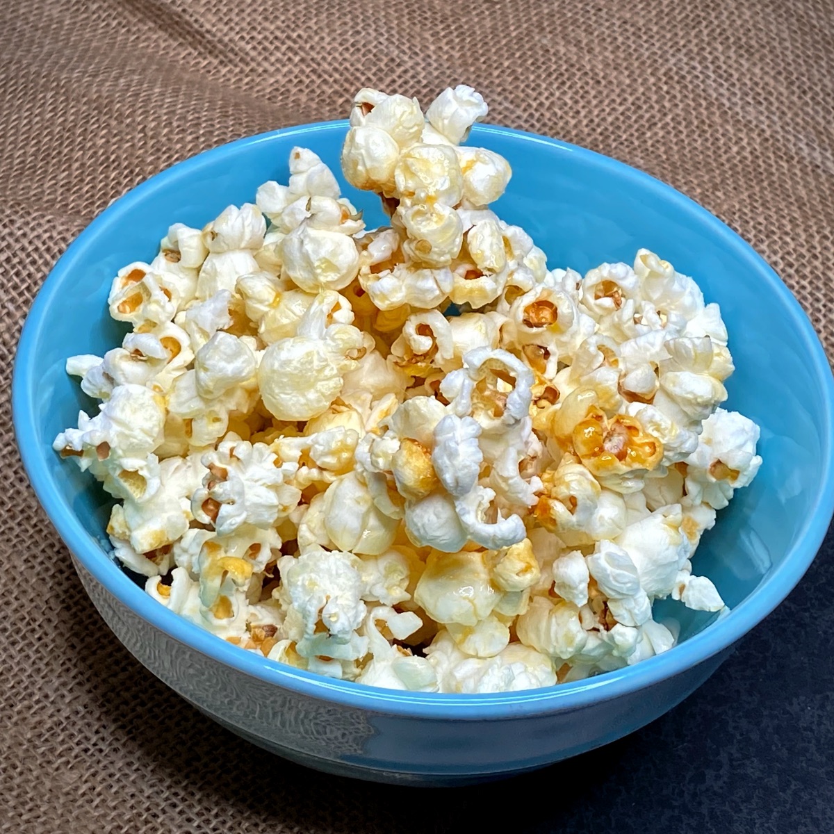 Top view of a bowl of homemade kettle corn in a blue bowl on a burlap table runner.