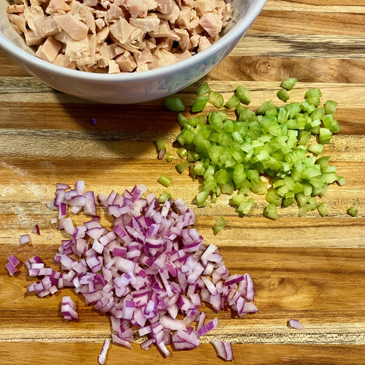 Overhead view of finely diced celery and red onion on a cutting board. A white bowl with cut up chicken is also shown.