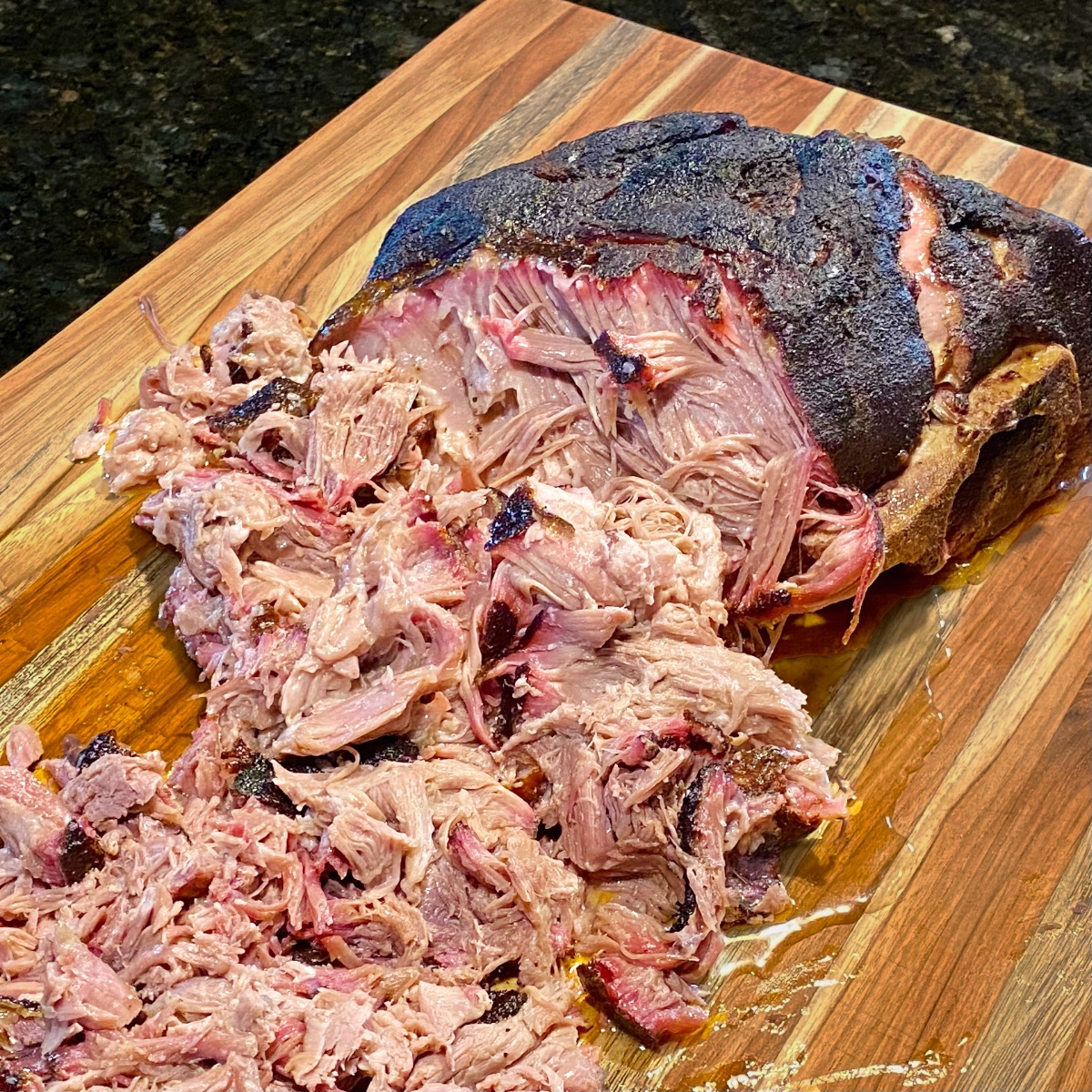 Smoked pork butt on a cutting board with half of it shredded (pulled). Image shows the smoke ring and how juicy it is.