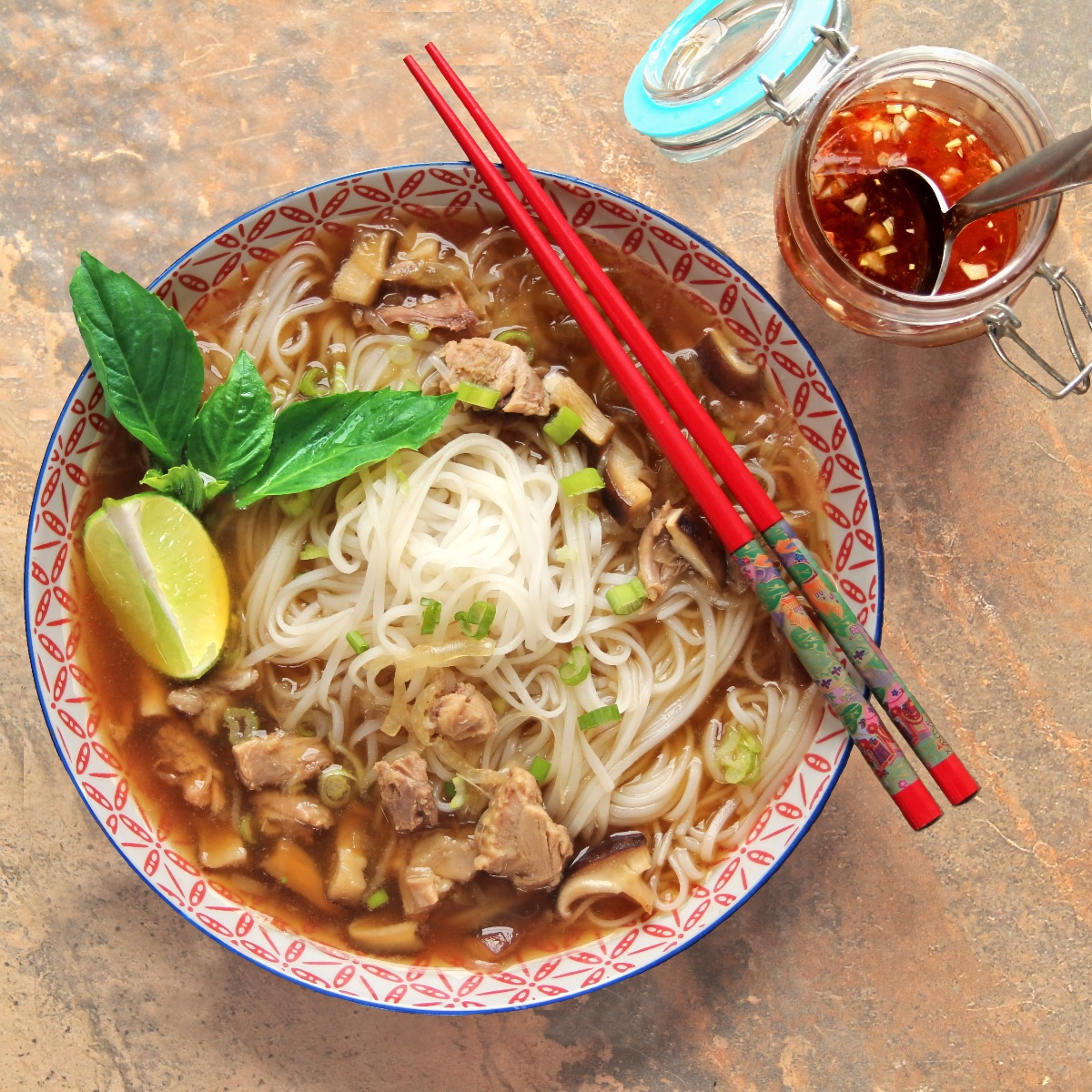 A serving of duck pho in a decorative red and white bowl with a set of red chopsticks laying on top. The pho has been garnished with a sprig of Thai basil and a lime wedge. A jar of nuoc cham sits nearby.