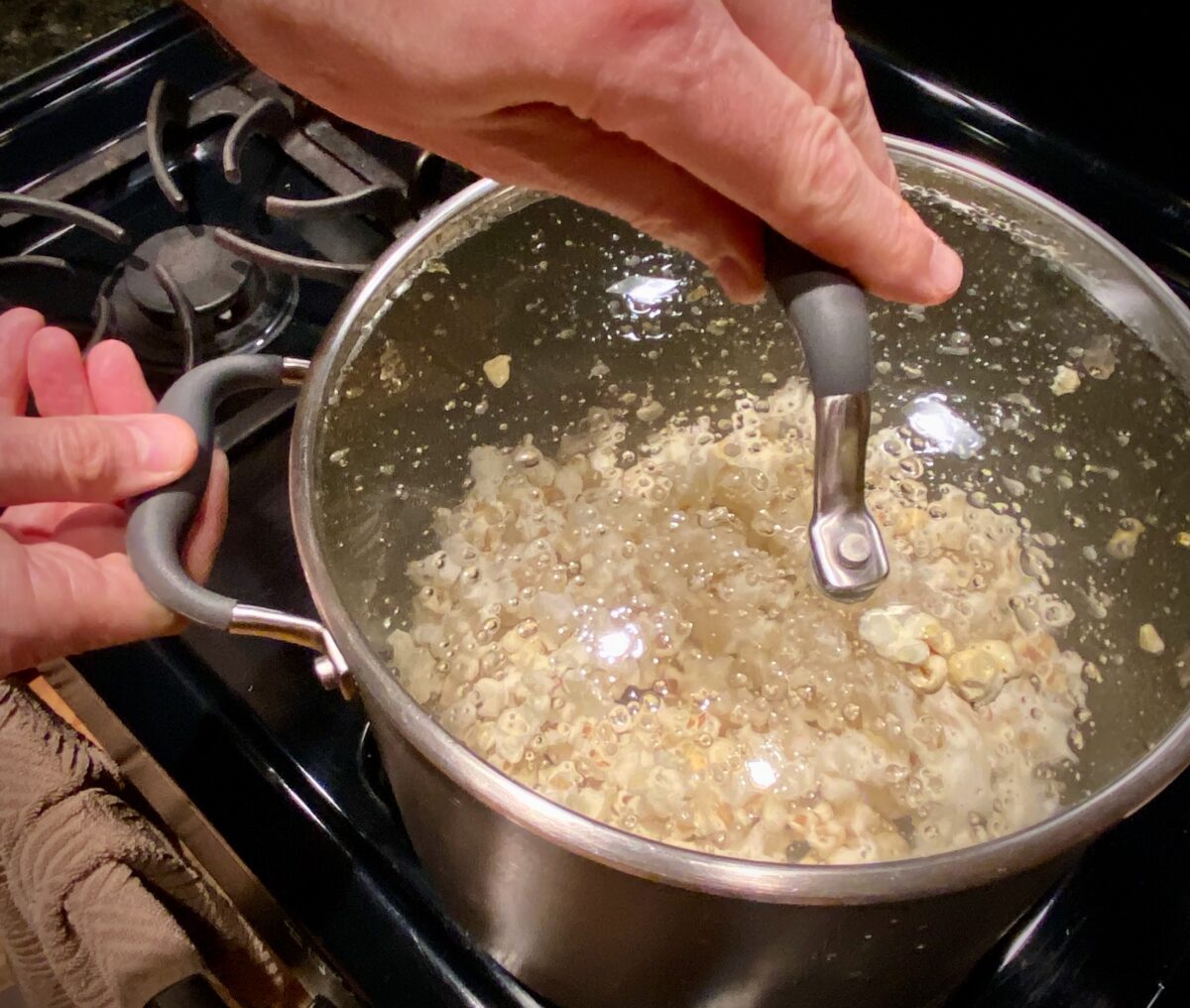 Top view of a person using one hand to hold the lid and the other hand to hold the pan while shaking the pan that has popcorn popping inside it.