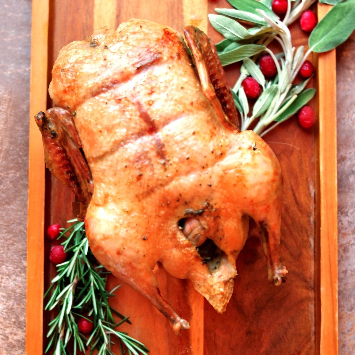 Finished Herbed Roast Duck on a wooden tray with cranberries and sage and roaemary sprigs.