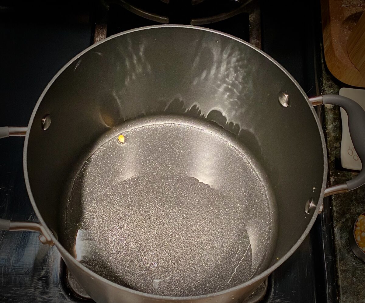 Top view of large pan with hot oil and one kernel of corn in it. The kernel is unpopped and at the far edge of the bottom of the pan. 